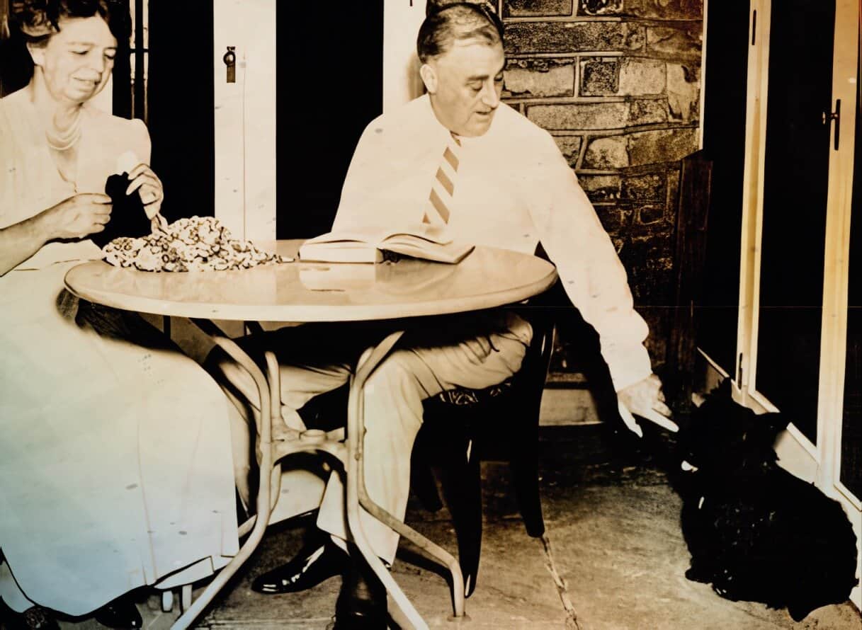 A vintage black-and-white photo of a man and woman at a table, with the woman opening a gift and the man looking down, capturing a nostalgic moment of mid-20th-century life.