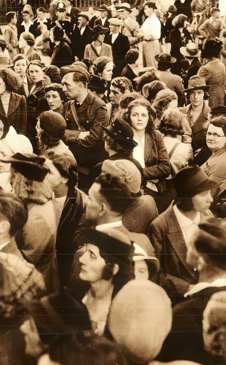 Crowd of men, women, and children dressed in early 20th-century fashion gathered outdoors, showing social diversity, with some engaging in conversation and others observing surroundings.