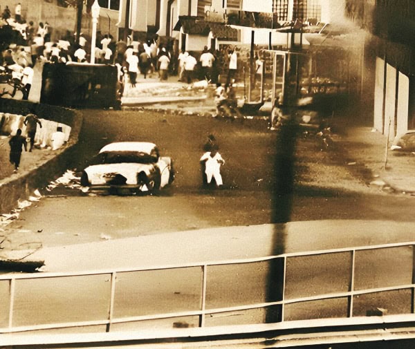 Burned-out cars and people gathered at a street scene during a protest or riot, capturing a moment of historical unrest and urban chaos, with a focus on civil disturbance and public demonstrations.