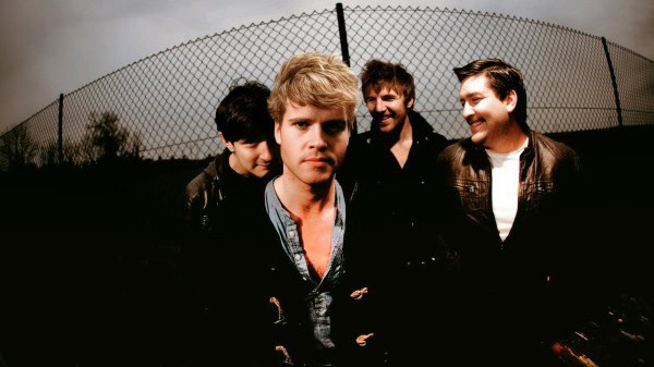 Young band members standing outdoors near a chain-link fence, showcasing a classic rock or indie style look for music event or promotion.
