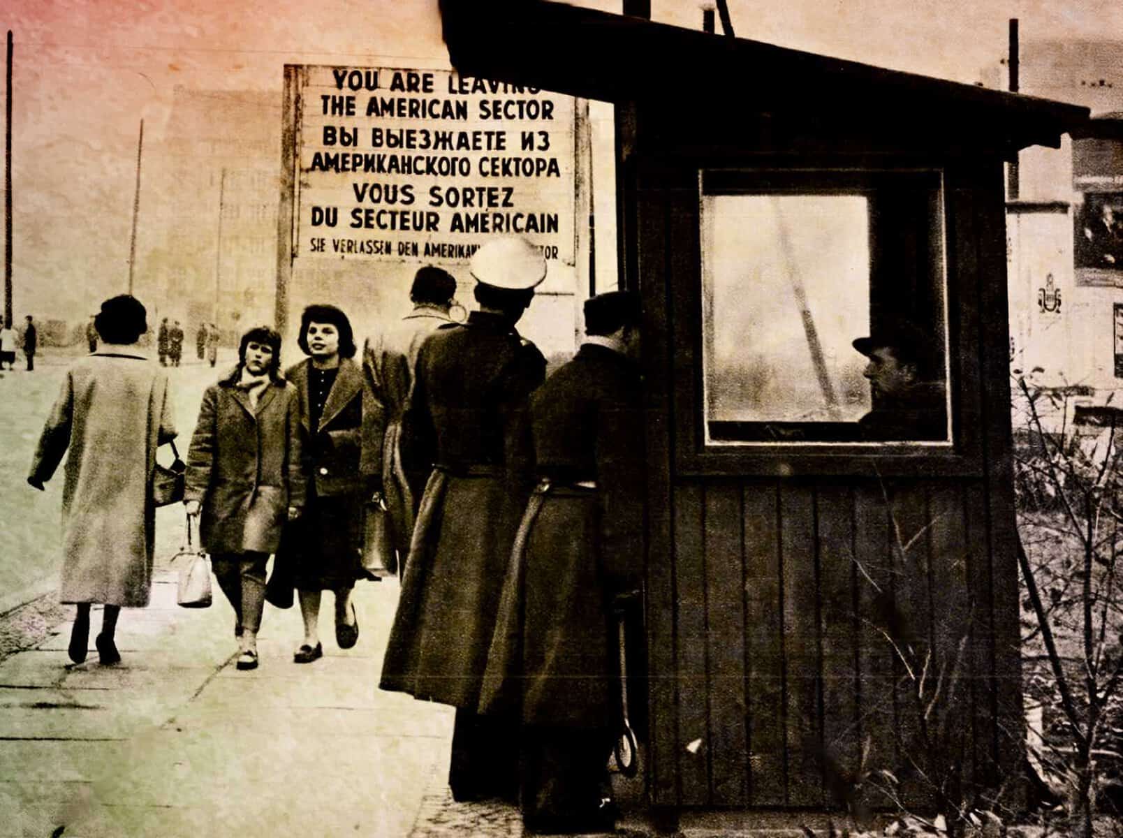 Historic black and white photo of people walking past a checkpoint with a sign about leaving the American sector, capturing moments of Cold War era history and urban life.