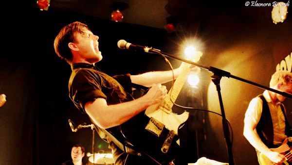 Guitarist performing live on stage during a music event, with bright stage lighting and an enthusiastic atmosphere.