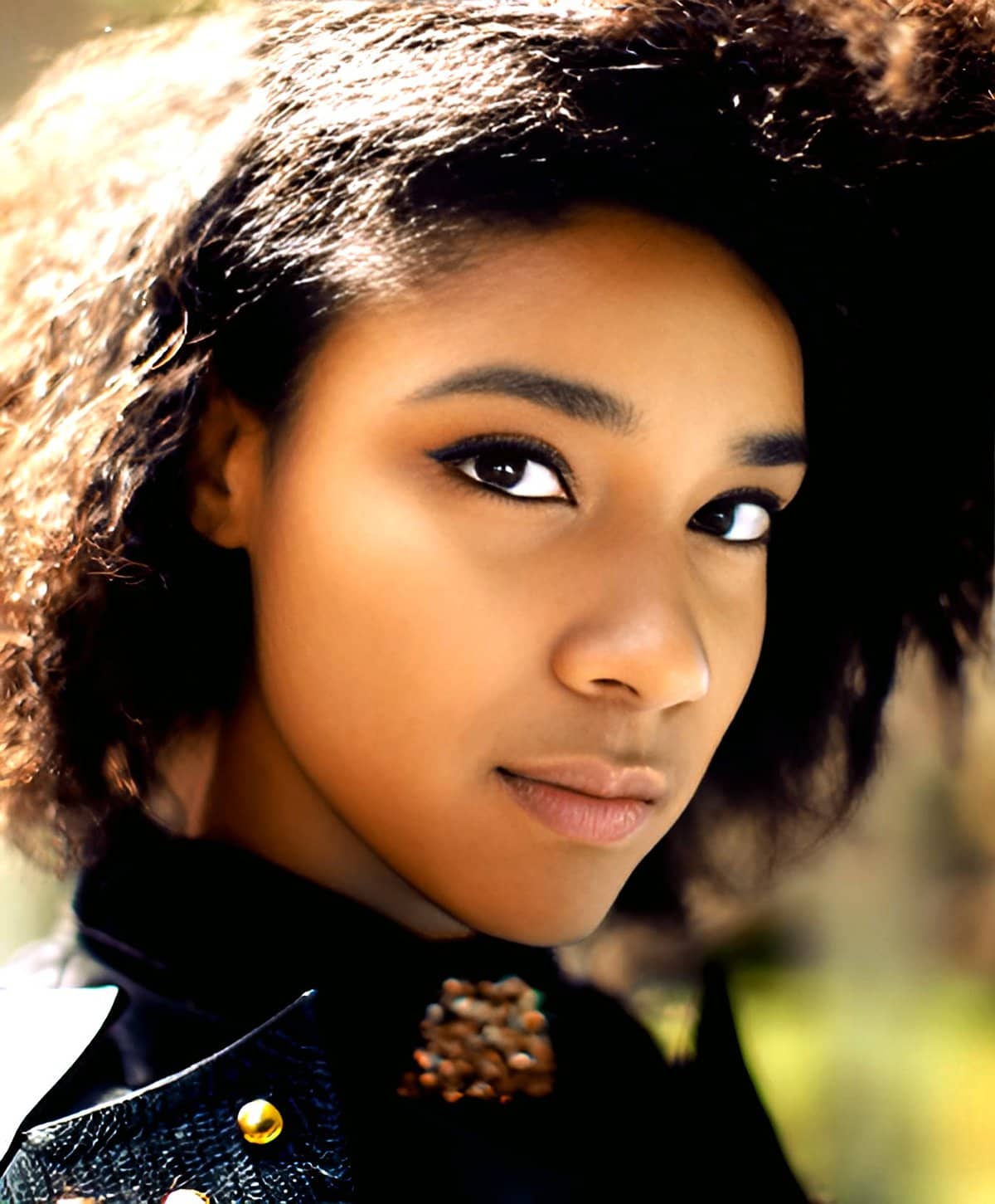 Portrait of a young woman with natural curly hair and subtle makeup, emphasizing her eyes, suitable for music, fashion, or lifestyle themes on a vibrant outdoor background.