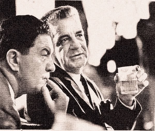 A vintage black and white photo of two men in conversation at a social gathering, with one man holding a glass of beer, capturing a historic moment in news, history, and music archives.
