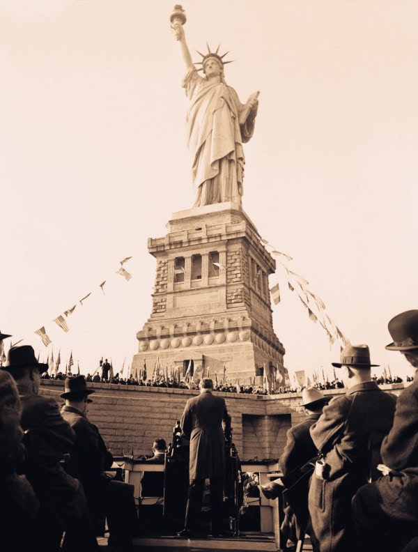 Liberty Statue in New York City during a historic event with people gathered at the base, capturing an iconic symbol of freedom and American history, featured in a vintage-style sepia tone for nostalgic appeal.