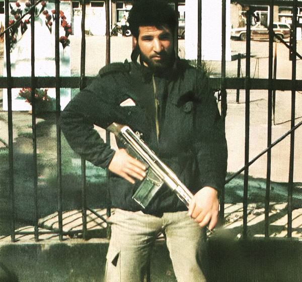 AK-47 assault rifle held by man in front of metal gate, urban background, vintage style photo.