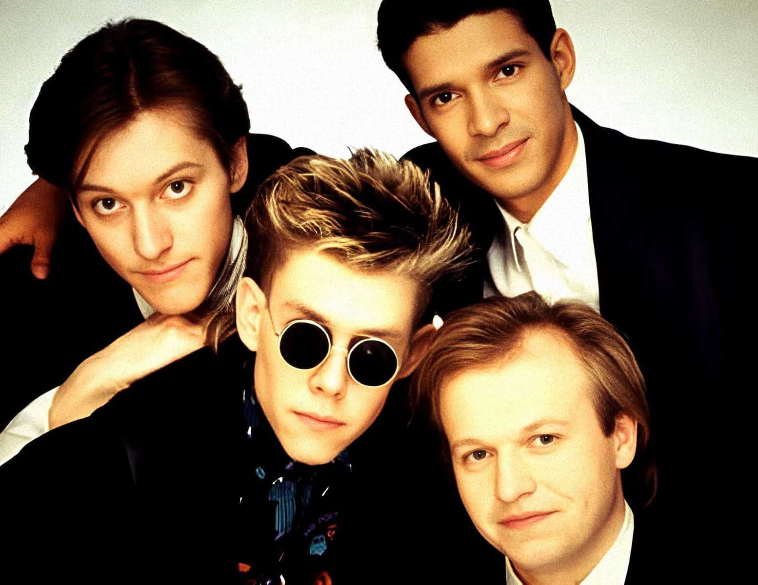 Young men in stylish clothing, some with sunglasses, posing closely together in a music or pop band photoshoot.