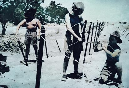 Historic black and white photograph of children playing outdoors, wearing hats, near a rustic wooden fence on a sunny day, capturing a nostalgic moment.