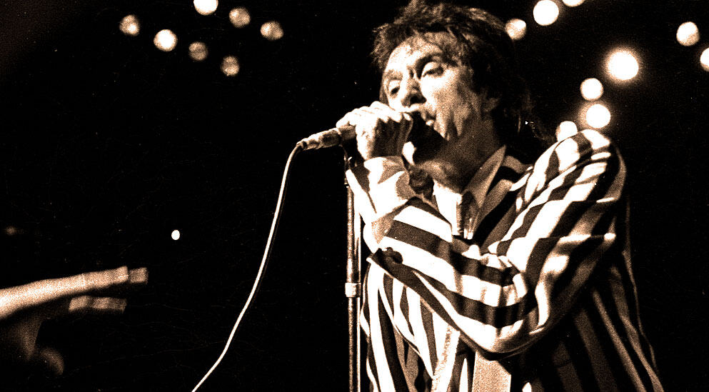 Retro black and white photo of a rock singer performing on stage with microphone, capturing vintage music performance style, emphasizing iconic musicians, history, and music archives.