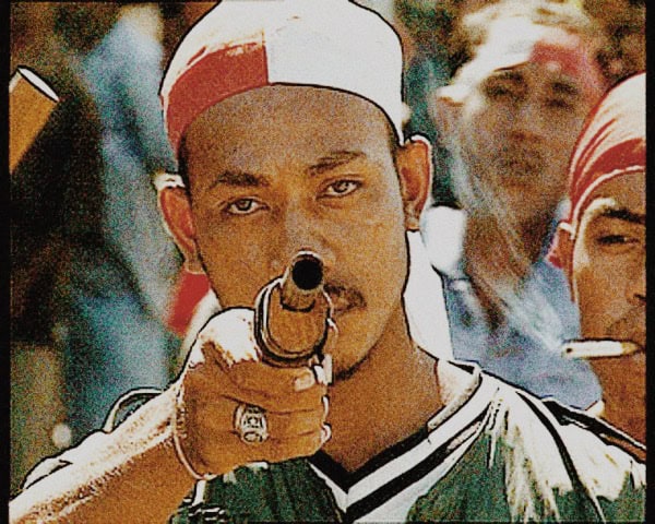 A young man in sports attire aiming a pistol directly at the camera, with a focused expression and a crowd in the background, capturing a moment of intense competition or protest.
