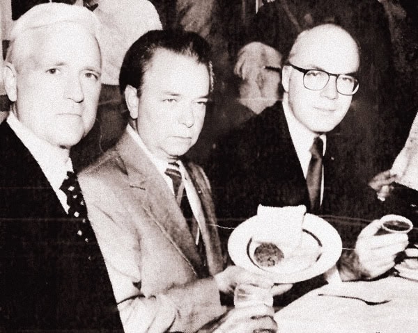 A black and white photo of three men in formal attire dining at a restaurant or event, capturing a historical moment related to news, music, or cultural history.