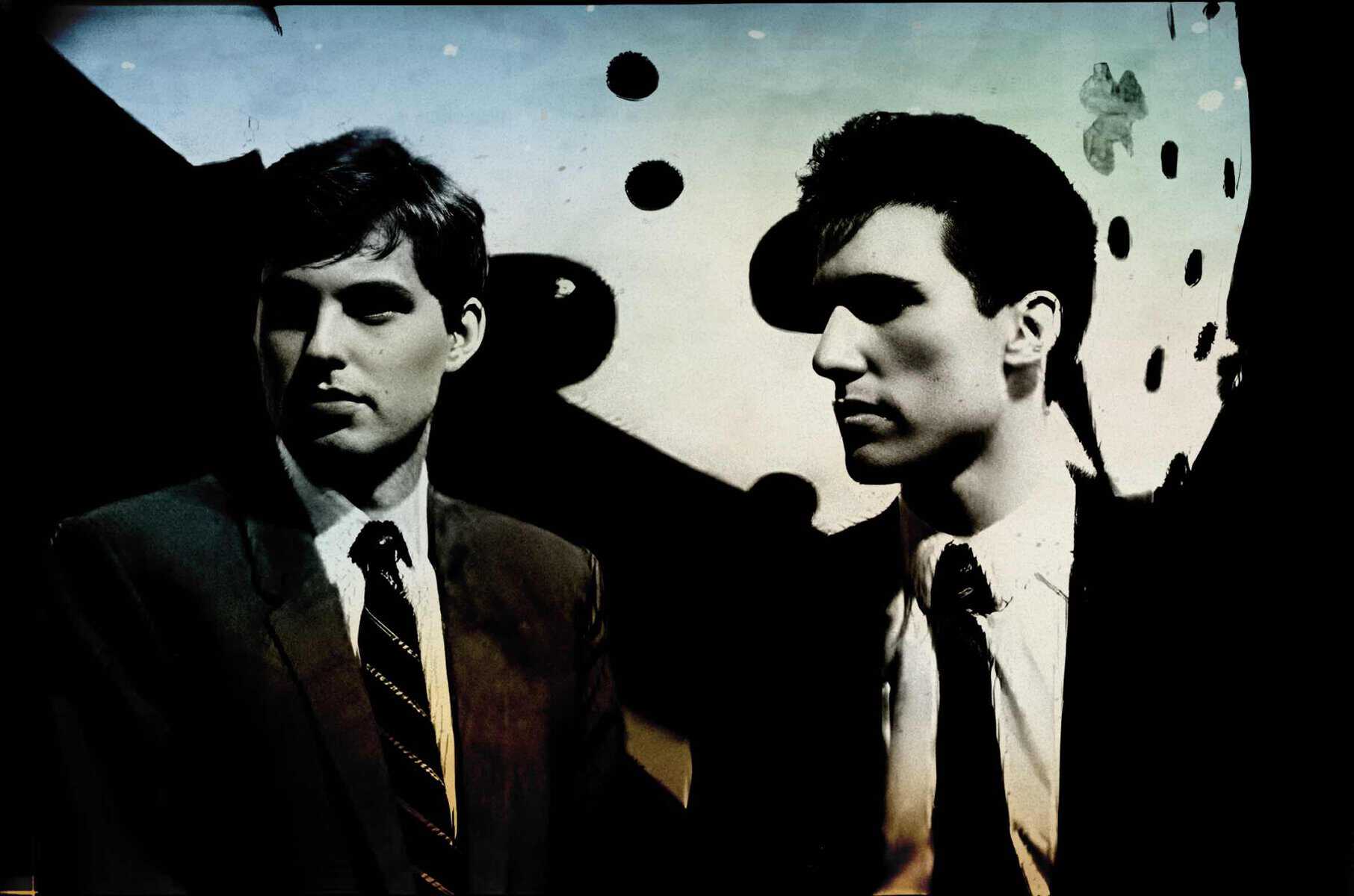 Vintage black and white photo of two young men in formal suits and ties, standing against an abstract painted background with circular shapes, capturing a retro music or news scene.