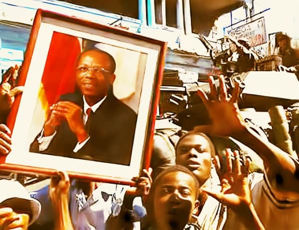 Serene man in formal attire holding framed photo at historic protest exploring music, news, and cultural history.