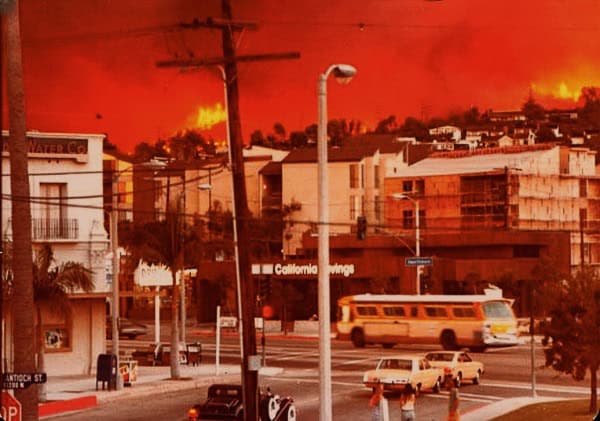Burning cityscape with fire and smoke over urban area, emergency scene with vehicles and traffic, intense orange sky, destruction and chaos, possibly related to wildfires or urban disaster, dramatic weather conditions, newsworthy event.