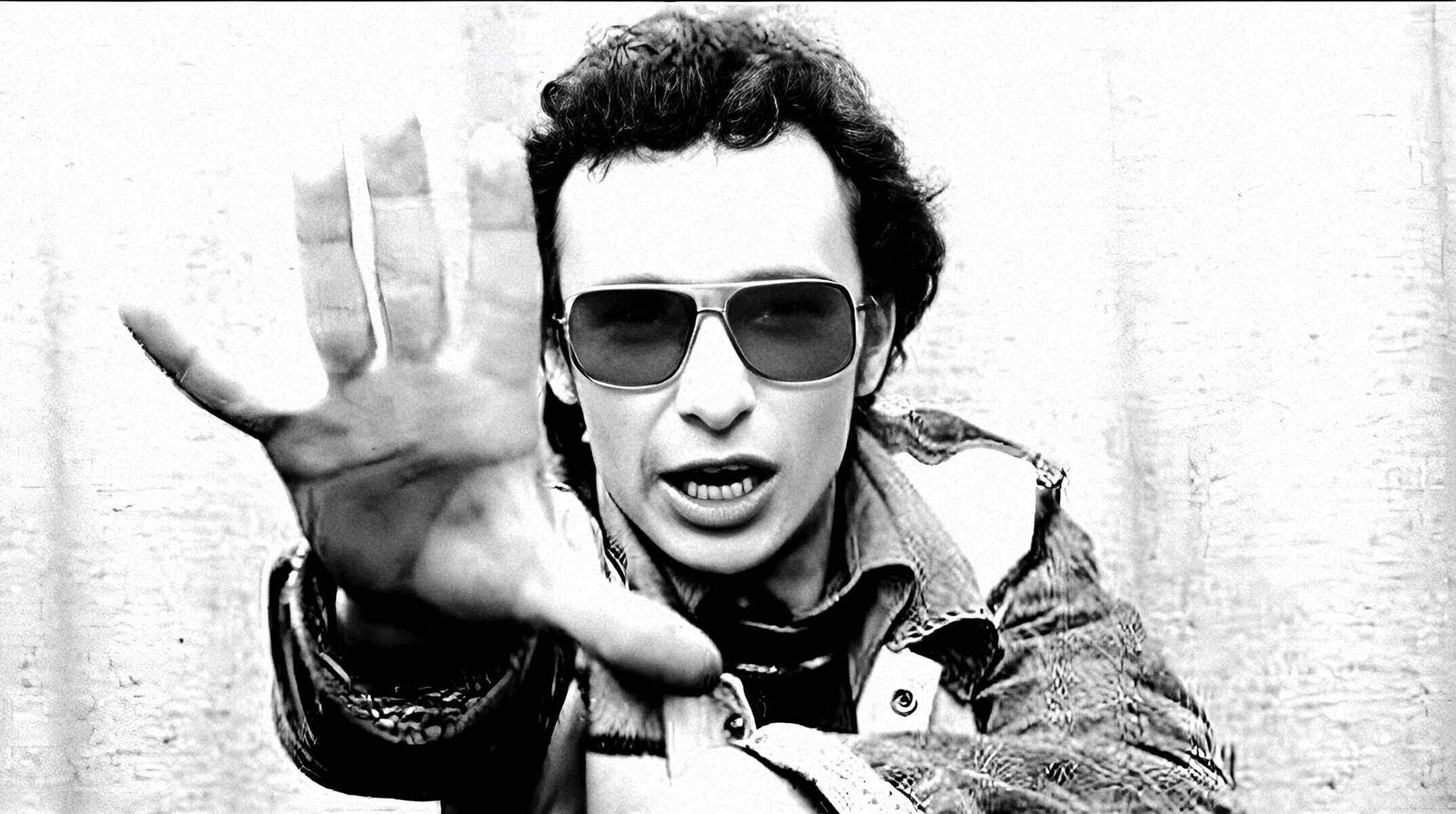 Close-up black and white image of a man in sunglasses making a stop gesture towards the camera, showcasing vintage fashion style, music, news archive content, and iconic moments in history.