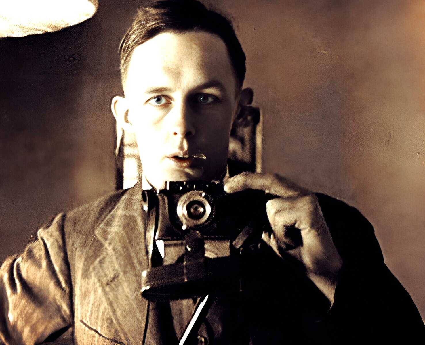 Old black and white photo of a young man with short hair holding a vintage camera to his face, capturing a photo. The image has a nostalgic aesthetic and emphasizes classic photography, history, and visual storytelling. |.