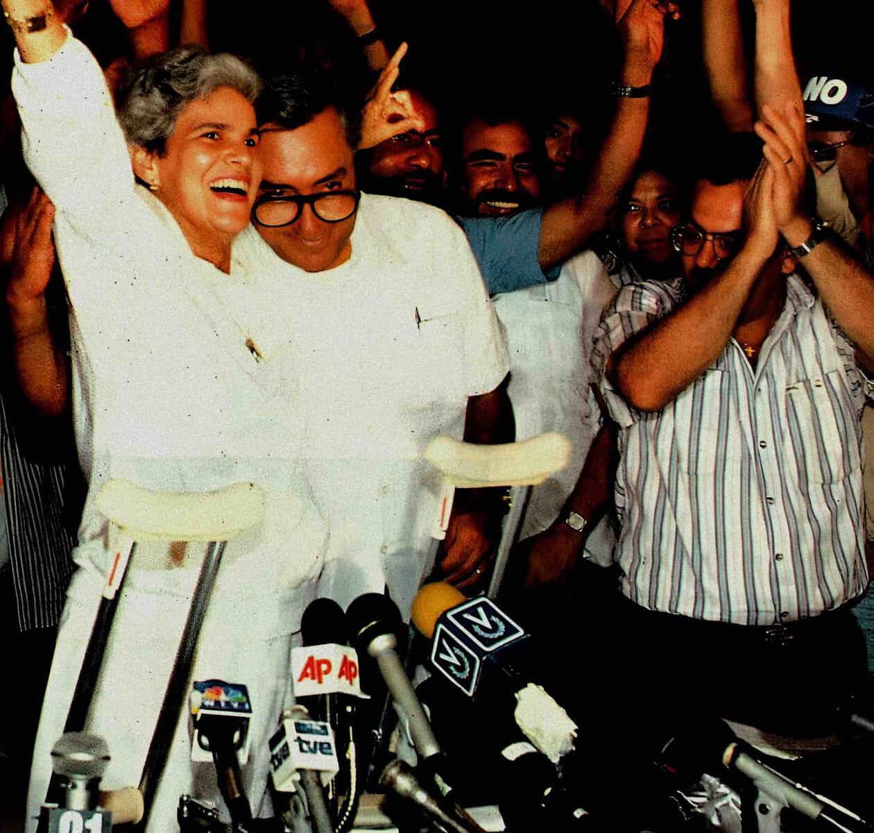 Disabled woman with crutches and white attire smiling at press conference, surrounded by journalists, in a lively news coverage scene.