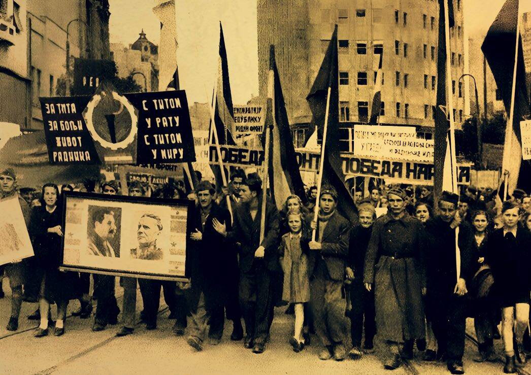 Historical black and white photograph of a march with people holding flags and banners, commemorating victory and historical events, related to news, history, and music archives.
