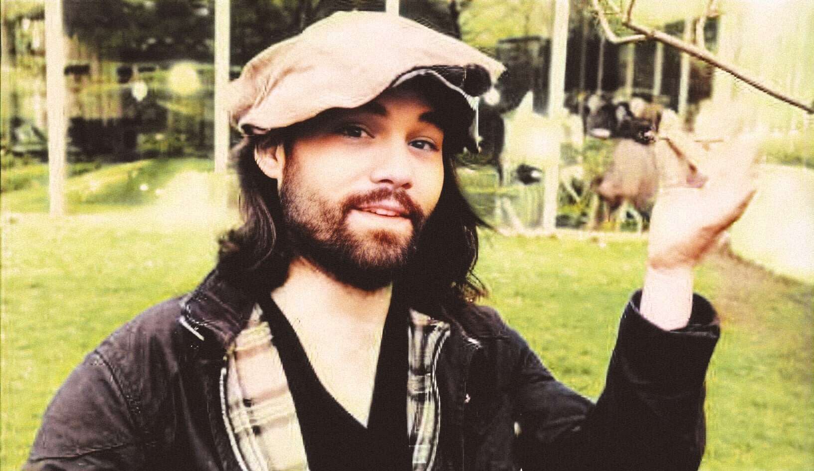 A man with long dark hair and beard, wearing a hat and plaid shirt, holding a fishing line with a small fish in an outdoor setting with green grass and trees.