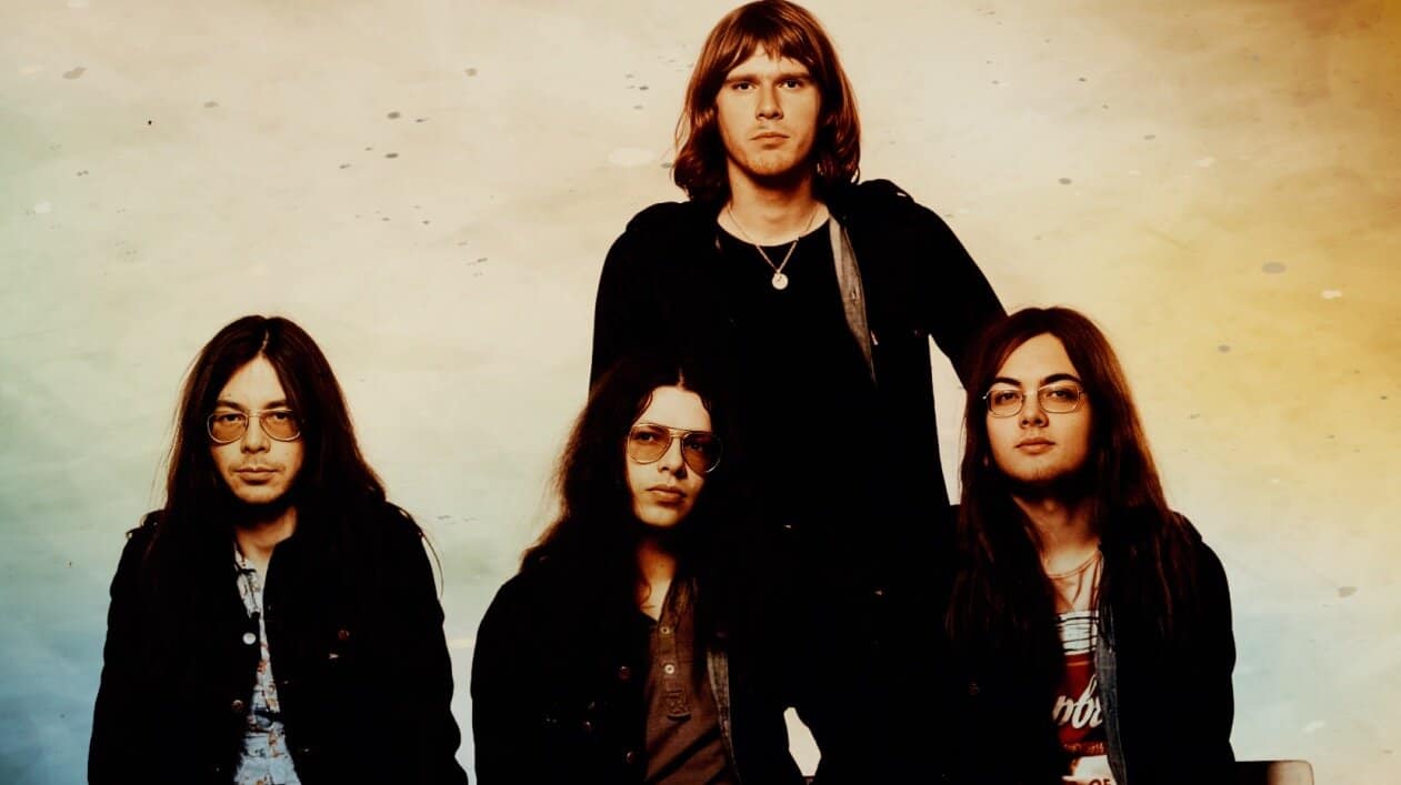 Vintage rock band portrait featuring four musicians with long hair, glasses, and casual dark clothing, representing music history and classic rock.