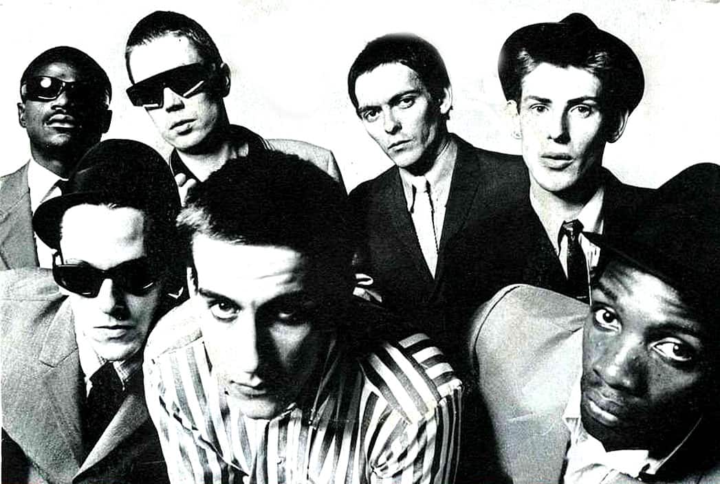 Black and white photo of a diverse 1980s music band with members wearing stylish sunglasses and suits, representing iconic musicians from the era, capturing vintage fashion and music history.