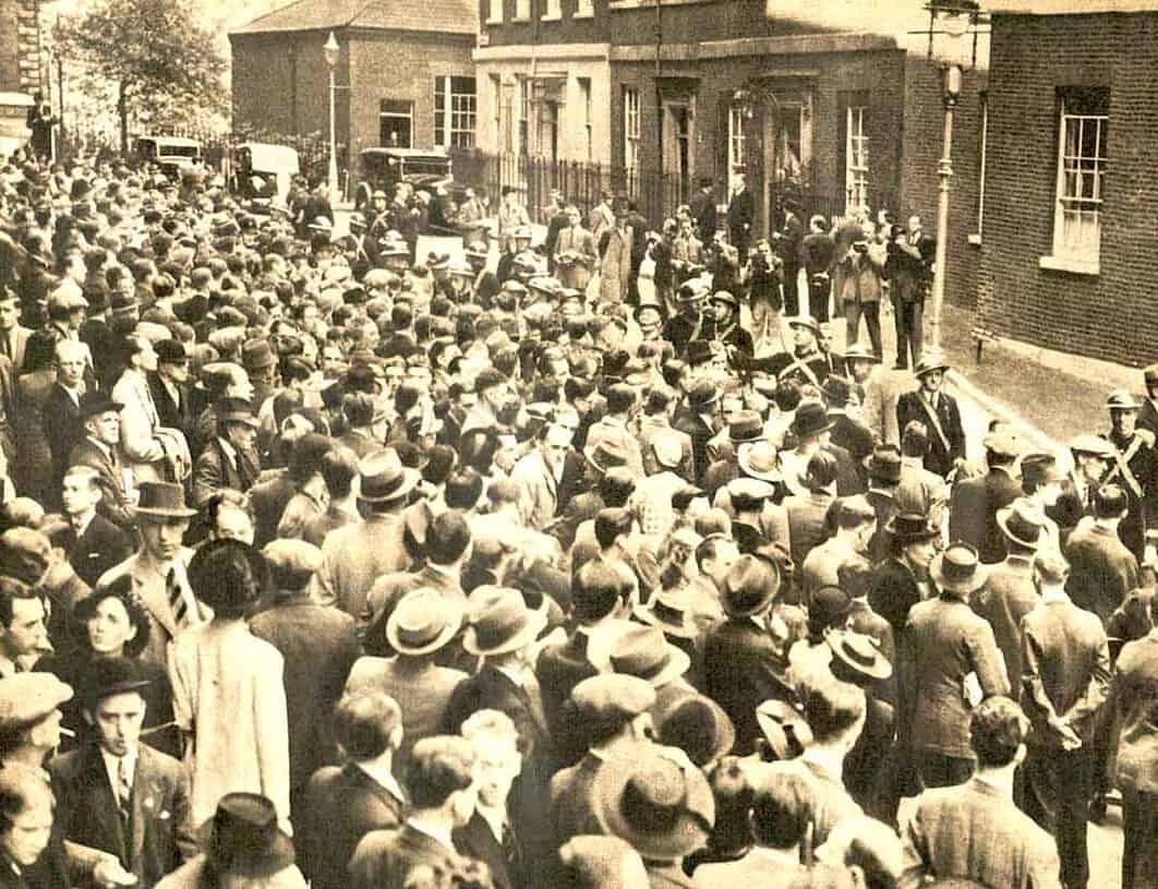 A large crowd of people gathered on a city street, many wearing hats, during an early 20th-century event or protest. The historic photo captures community engagement and urban life from that era.