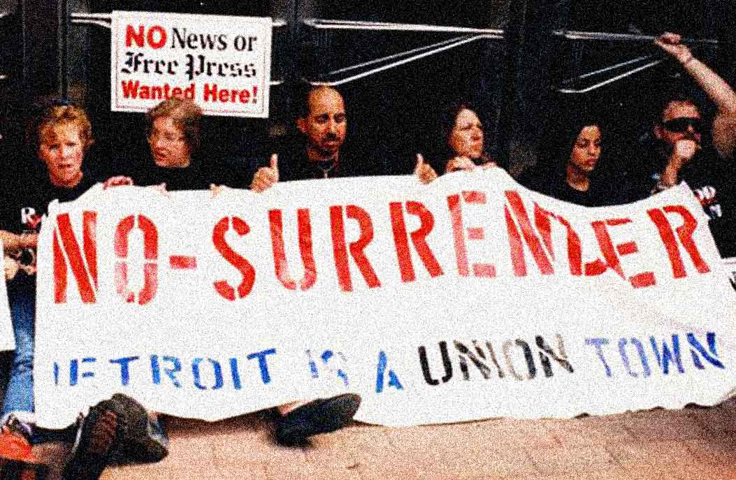 No-surender protest with women holding a large sign reading "NO-SURRENDER" against press repression, sign demanding "NO News or Free Press Wanted Here!", emphasizing press freedom and community activism.