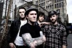 Band of four young men outdoors near urban buildings, street scene, tattooed member in center, casual grunge style, capturing alternative music culture.