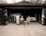 Children and adults sitting outside a garage filled with bicycles, vintage car, and gardening tools, capturing a nostalgic family moment.