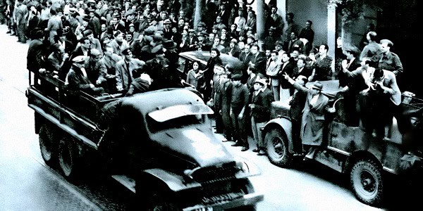 Black and white historic photo of a 1940s crowd scene with military trucks and civilians, capturing a moment of wartime activity and news coverage.