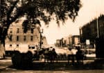 Vintage black-and-white photo of a historic downtown city street with people sitting on benches, old buildings, and a tree-shaded park, capturing the essence of vintage news, history, and music archives.
