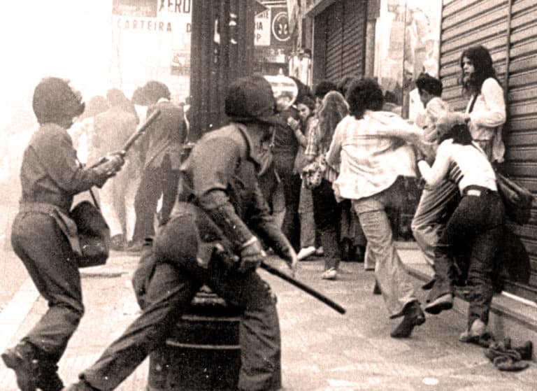 Riot police clash with protesters in urban street scene during civil unrest.