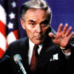 Portrait of a serious man in a dark pinstripe suit making a gesture with his hand during a press conference, American flag in background, emphasizing news, politics, and media history for SEO.