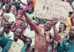 Protesters holding a sign "Welcome Home de MENDLA" during rally or demonstration, African community rally, news, history, music, protest history, social activism, cultural demonstration, historical sound archive, Past Daily: A Sound Archive of News, History And Music.
