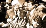 A vintage black and white photo of a group of politicians and journalists at a press conference with multiple microphones, capturing a historical moment in news, politics, and media history.