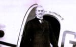 Vintage black and white photograph of a man with a mustache in early 20th-century attire standing at an airport entrance.