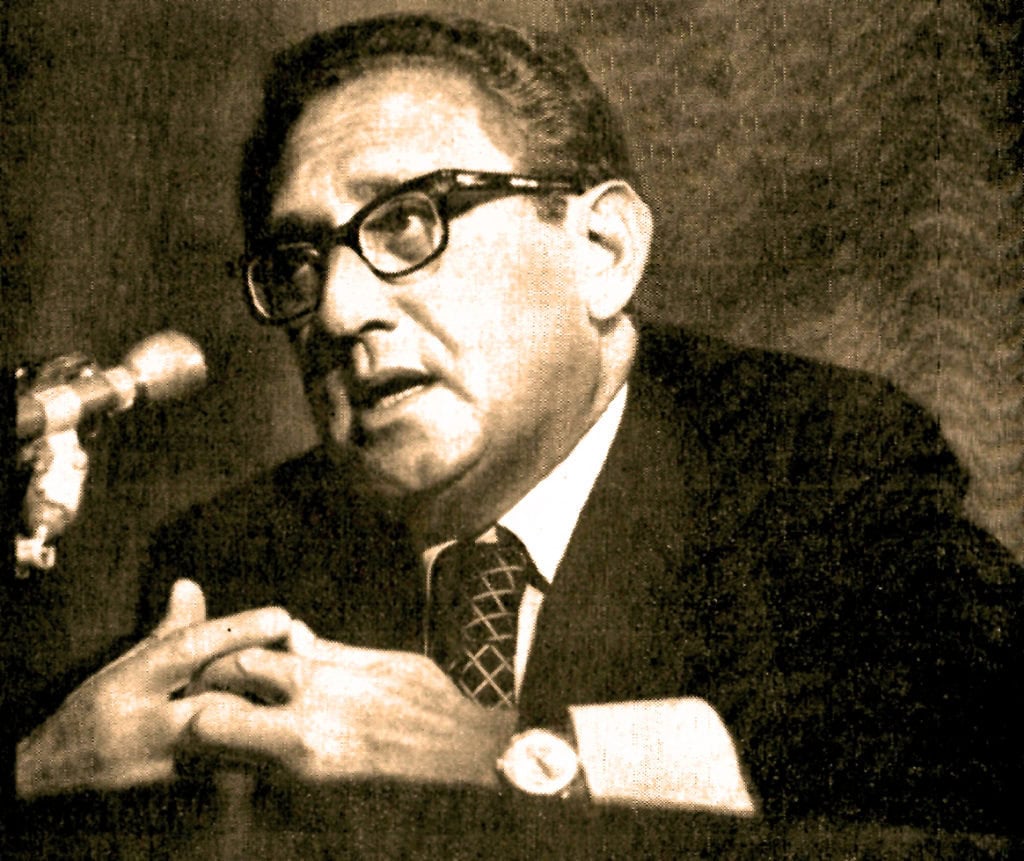 Historic black and white photograph of a man in glasses speaking into a microphone, capturing a moment of news or history documentation.
