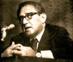 Historic black and white photograph of a man in glasses speaking into a microphone, capturing a moment of news or history documentation.
