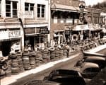 Vintage Main Street with tire stacks and local businesses, capturing mid-20th century small-town life with theaters, shops, and parked cars.