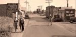 Mid-20th century small town street scene with vintage cars, local stores, and pedestrians, capturing historical Americana and community life from the era.
