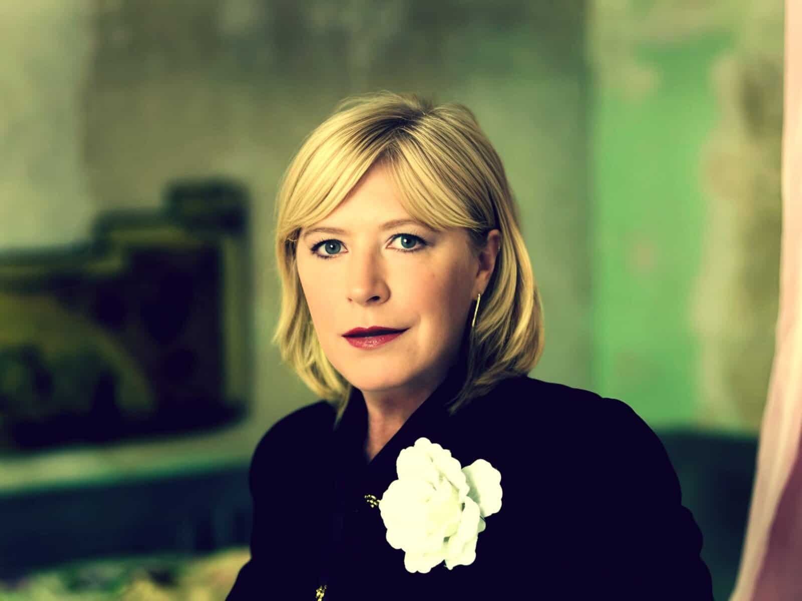 Elegant woman with blonde hair and blue eyes, wearing a black outfit and a white floral brooch, in a professional portrait setting.