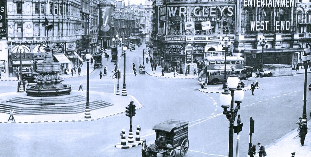 Vintage black and white image of a busy early 20th-century city street with a fountain, horse-drawn carriage, double-decker bus, and pedestrians, capturing urban life and entertainment signs.