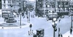 Vintage black and white image of a busy early 20th-century city street with a fountain, horse-drawn carriage, double-decker bus, and pedestrians, capturing urban life and entertainment signs.