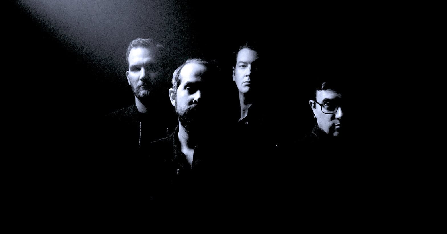 Dark portrait of a band with four male members, emphasizing shadows and serious expressions, representing music archives, news, and history at Past Daily.