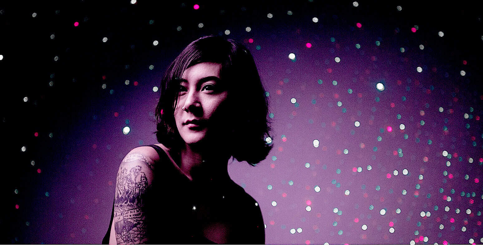 Vibrant portrait of a young woman with tattoos and dark hair, surrounded by colorful bokeh lights, illustrating music, history, and news archives for SEO on Past Daily Sound Archive.