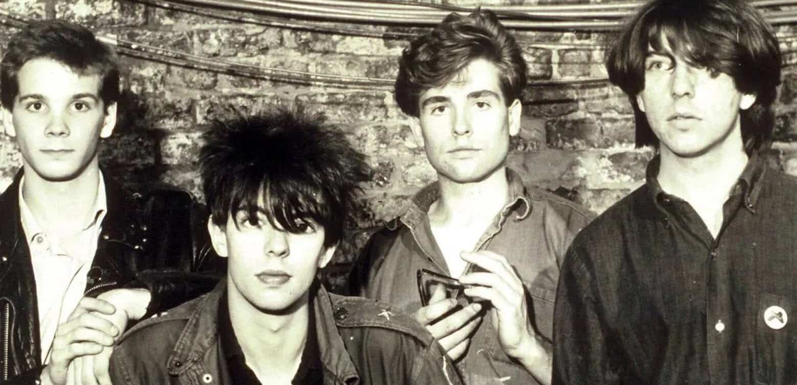 Black and white photo of a young alternative rock band with four members, standing against a brick wall, with one member holding glasses, capturing 1980s music scene.