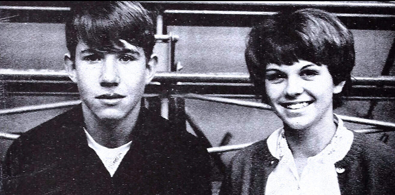 Youthful man and woman in black and white photo, smiling and looking at the camera, vintage setting with seats and railings in background, capturing a nostalgic moment from past history.