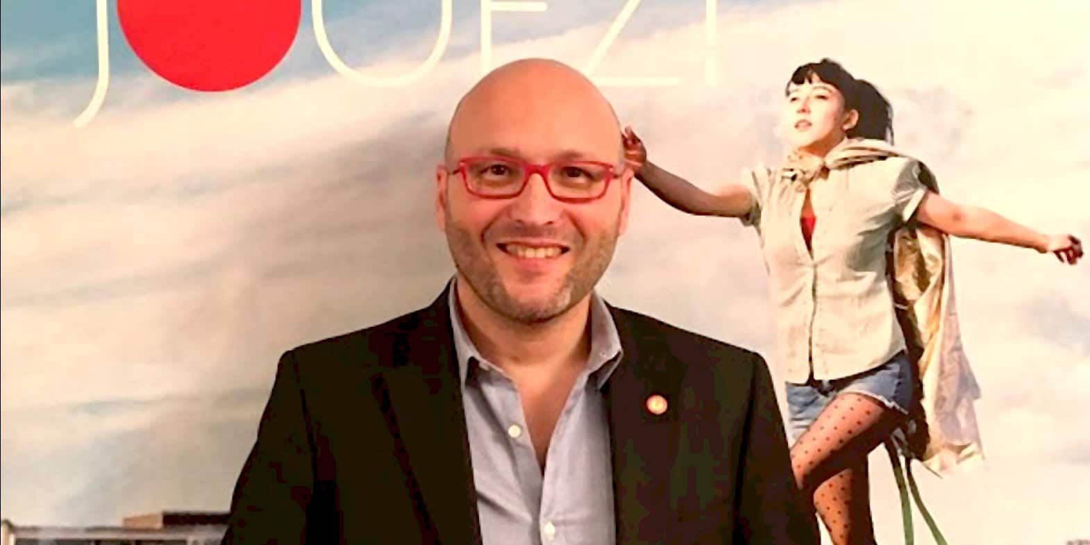Bald man with glasses smiling in front of a colorful background featuring an artistic woman image, representing historical news and music archive at Past Daily.