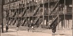 An old black and white photo of a fire escape on a brick building with women and children standing outside, historic urban scene, vintage city life, nostalgia, early 20th century architecture.