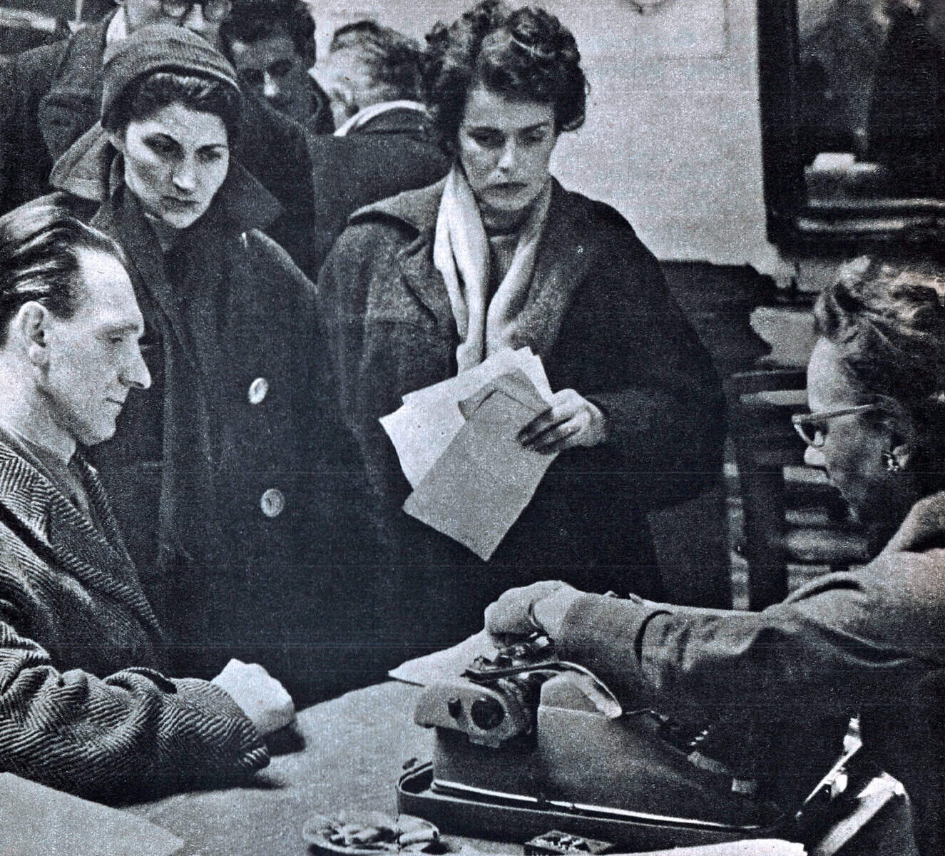Old black and white photo of women and men in a vintage newsroom or radio station, engaged in news reporting or listening to broadcasts, capturing mid-20th century media history for historical archives.